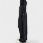 Asymmetrische Hose in Schwarz mit weitem Bein und seitlicher Naht, Knopfverschluss sichtbar.