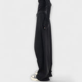 Asymmetrische Hose in Schwarz mit weitem Bein und seitlicher Naht, Knopfverschluss sichtbar.