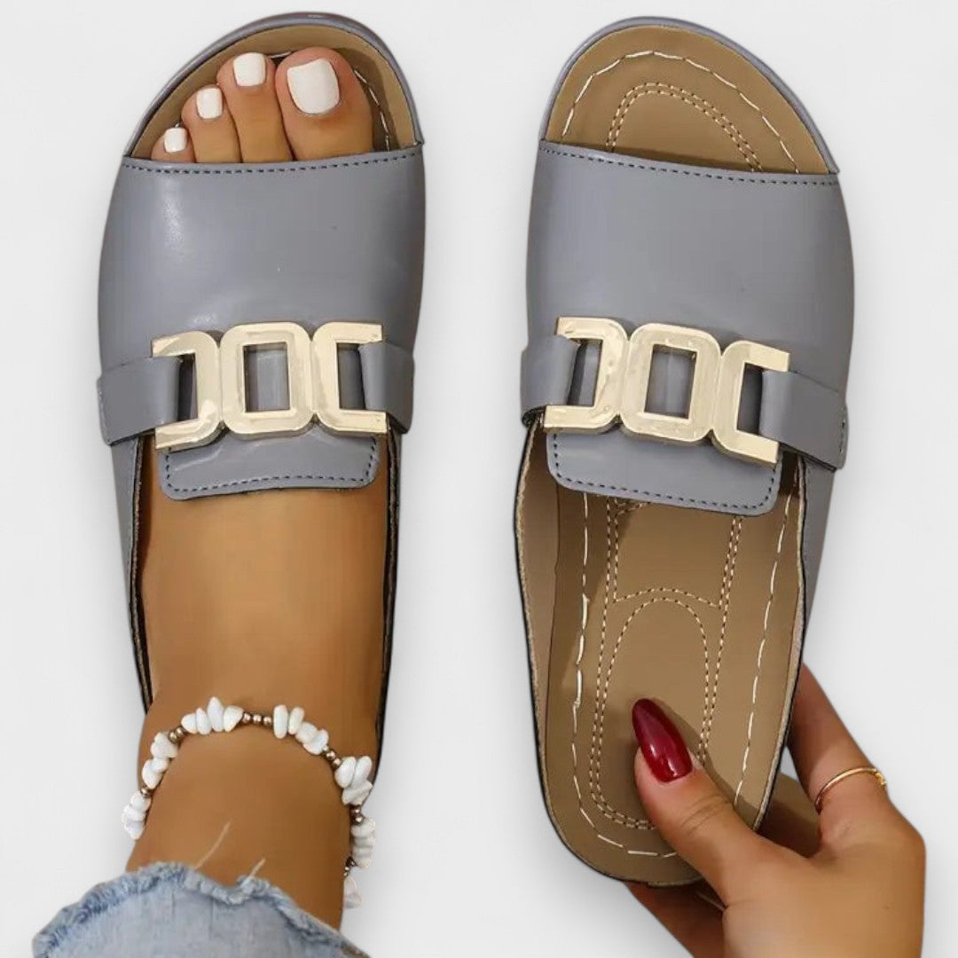 Graue Sandalen mit breitem Riemen und goldfarbenem Logo-Element, offene Zehenpartie